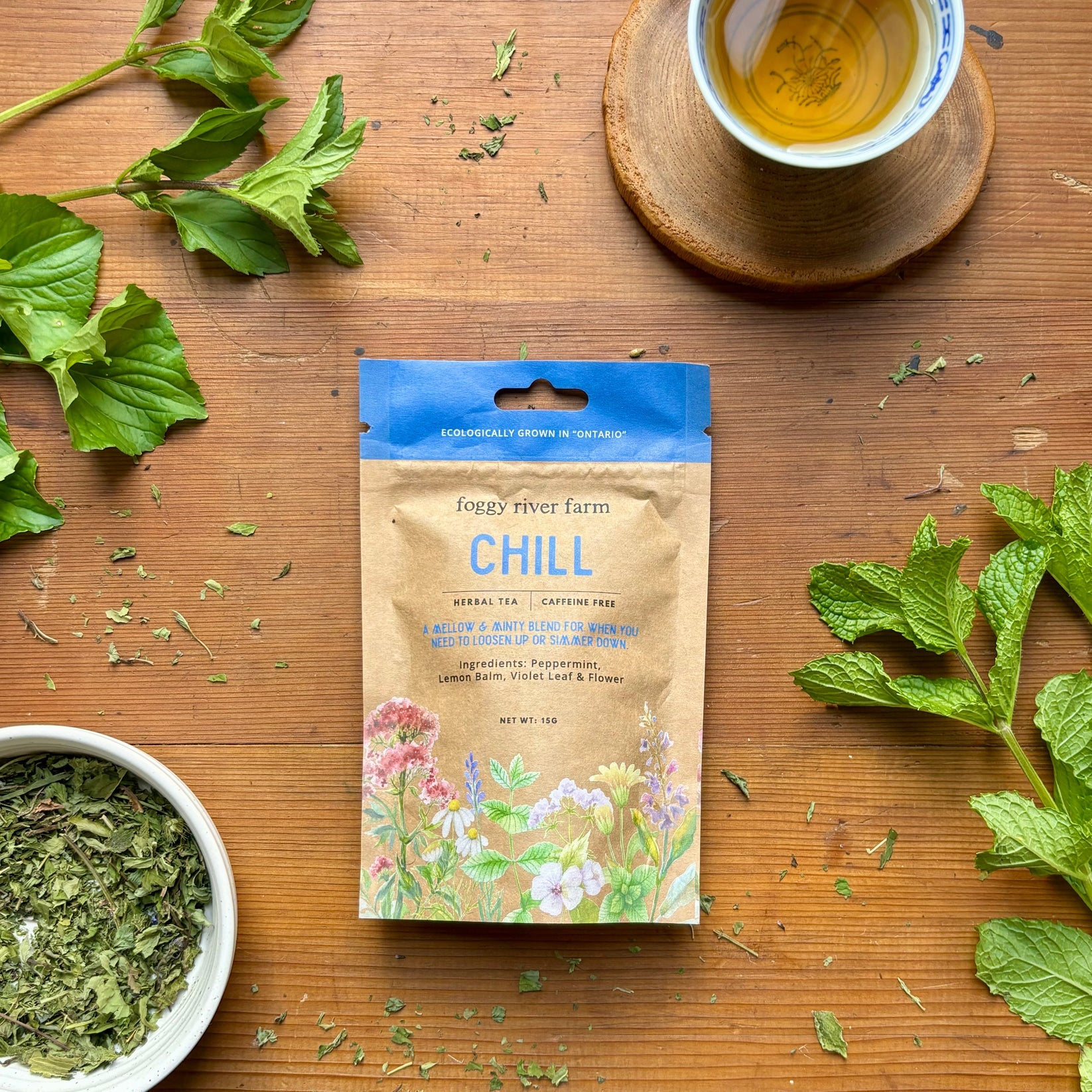 Tea package labeled 'Chill' by Foggy River Farm on a wooden surface with tea leaves and a cup of tea.